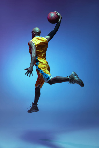 Handsome african-american male basketball player in action and motion in neon light on blue background. Concept of healthy lifestyle, professional sport. - Foto, Imagem