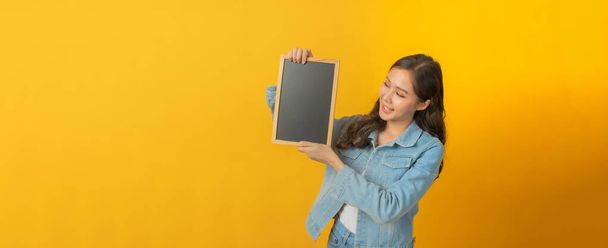 panoramic banner. happy asian pretty woman in casual clothing smile, hold empty blank blackboard with copy space area for text on yellow background, online marketing, advertising, announcement concept - 写真・画像