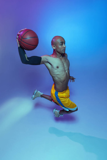 Sportive athletic african-american male basketball player posing in neon light on blue background. - Φωτογραφία, εικόνα