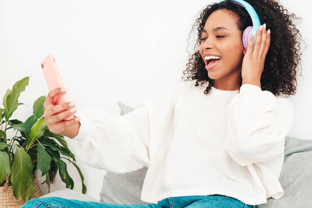 Beautiful black woman with afro curls hairstyle.Smiling model in sweater and jeans.Sexy carefree female listening music in wireless headphones in morning.Lying in bed in white interior.Taking selfie - 写真・画像