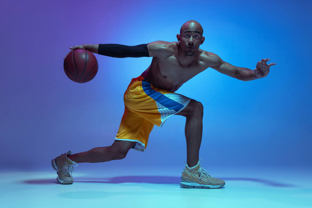 Sportive athletic african-american male basketball player training in neon light on blue background. - Φωτογραφία, εικόνα