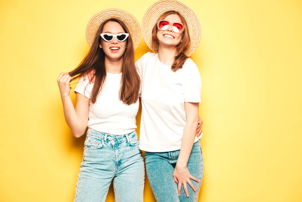 Two young beautiful smiling hipster female in trendy summer white t-shirt and jeans clothes.Sexy carefree women posing near yellow wall in studio.Positive models in hats - Valokuva, kuva