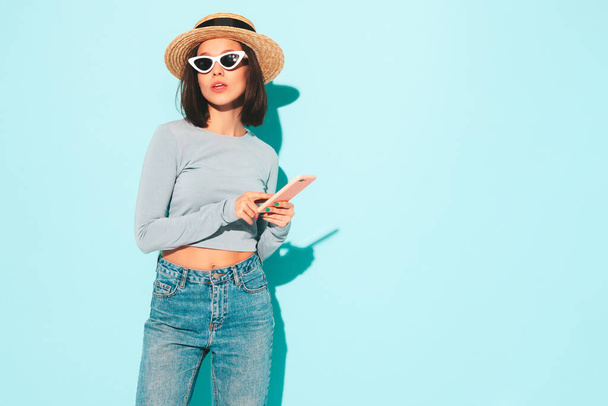 Young beautiful smiling female in trendy summer hipster  clothes. Sexy carefree woman posing near blue wall in studio. Positive model having fun in sunglasses and hat. Holds smartphone, using apps - Foto, afbeelding