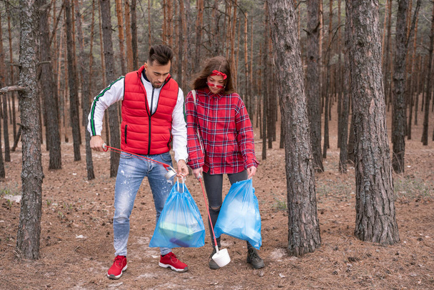 νεαρό ζευγάρι με σακούλες σκουπιδιών μαζεύοντας σκουπίδια με εργαλεία αρπαγής στο δάσος - Φωτογραφία, εικόνα