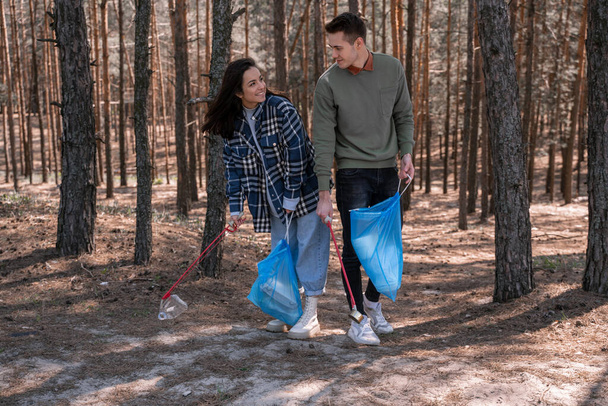 happy young woman and man with trash bags picking up rubbish with grabber tools in forest  - Photo, Image