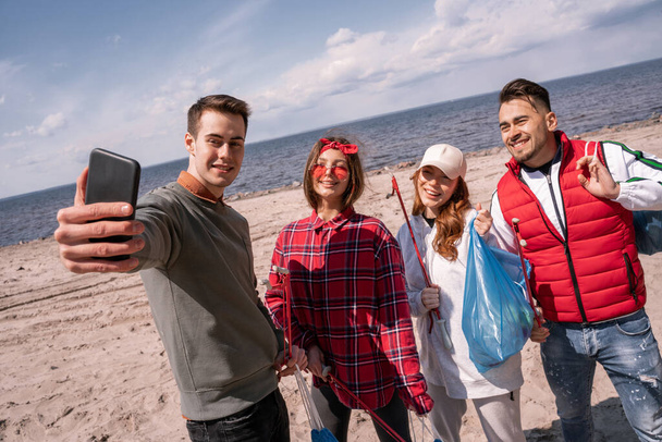 felice giovane uomo prendendo selfie con gli amici mentre raccogliendo spazzatura  - Foto, immagini