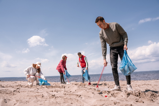 ευτυχισμένος άνθρωπος κρατώντας σακούλες σκουπιδιών και μαζεύοντας σκουπίδια στην άμμο κοντά σε μια ομάδα εθελοντών  - Φωτογραφία, εικόνα