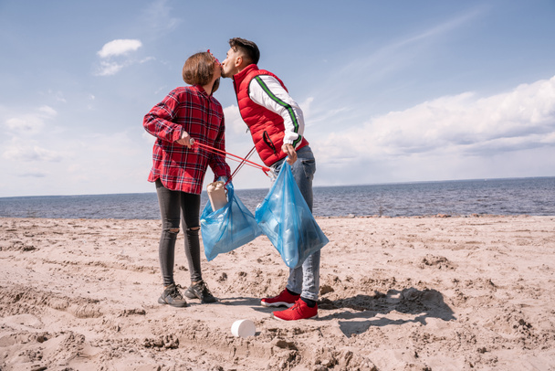 coppia con sacchetti della spazzatura e afferratori baciare mentre in piedi sulla sabbia  - Foto, immagini