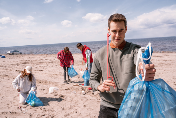 uomo felice tenendo sacchetto della spazzatura vicino a un gruppo di volontari offuscati raccogliendo spazzatura sulla sabbia  - Foto, immagini