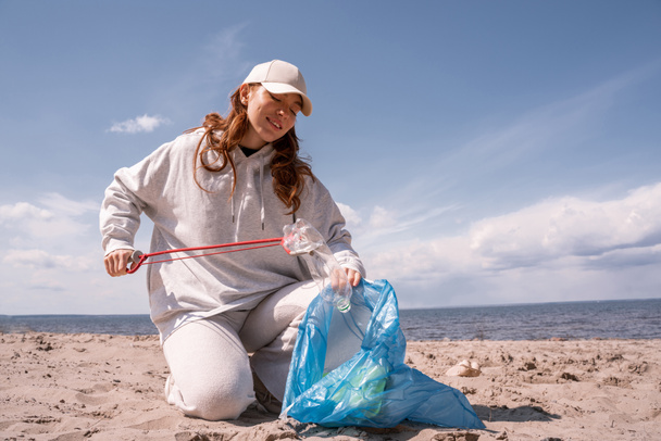 donna felice in tappo tenendo sacchetto della spazzatura e raccogliendo spazzatura sulla sabbia  - Foto, immagini
