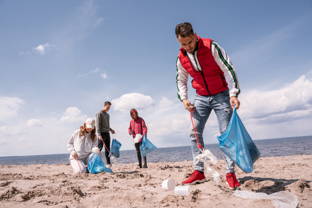 giovane uomo tenendo sacchetto della spazzatura e raccogliendo rifiuti sulla sabbia vicino ai volontari  - Foto, immagini