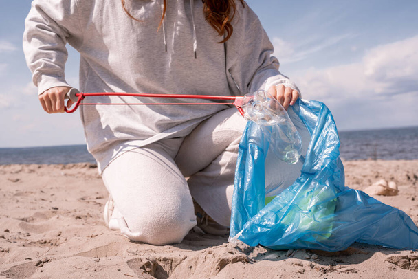vista ritagliata della donna tenendo sacchetto della spazzatura e raccogliendo spazzatura sulla sabbia  - Foto, immagini