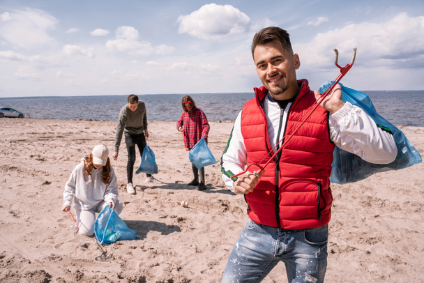 uomo allegro tenendo sacchetto della spazzatura e afferrare vicino volontari offuscati raccogliendo spazzatura sulla sabbia  - Foto, immagini