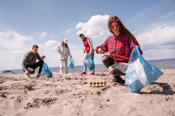 χαμογελαστή νεαρή γυναίκα που κρατάει σακούλες σκουπιδιών και μαζεύει σκουπίδια με αρπαγή άμμου κοντά σε ομάδα εθελοντών  - Φωτογραφία, εικόνα