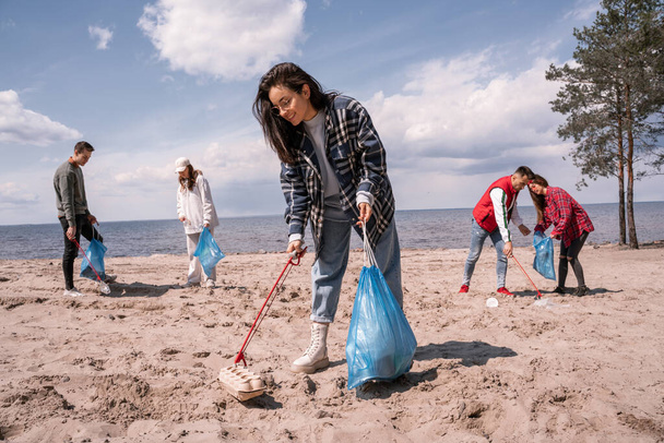 giovane donna sorridente tenendo sacchetto della spazzatura e raccogliendo spazzatura sulla sabbia vicino a un gruppo di volontari  - Foto, immagini