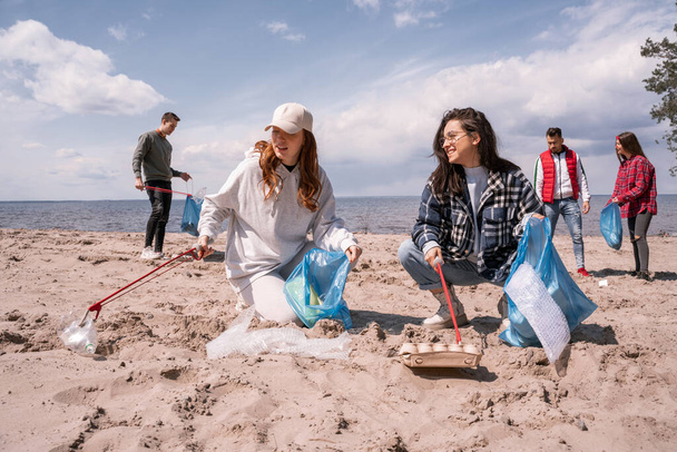 giovani donne sorridenti che tengono sacchi della spazzatura e raccolgono rifiuti con afferratori sulla sabbia vicino a un gruppo di volontari  - Foto, immagini