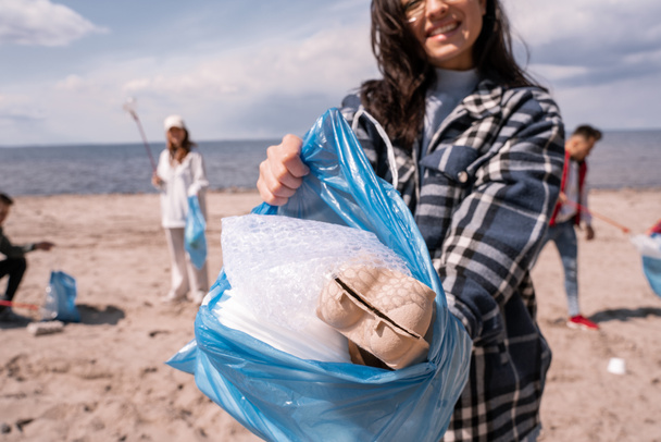 vista ritagliata di giovane donna sorridente tenendo sacchetto della spazzatura con spazzatura vicino gruppo di volontari  - Foto, immagini