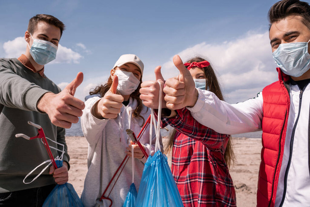 giovani volontari in maschere mediche in possesso di sacchetti della spazzatura e afferratori, mostrando i pollici verso l'alto, raccogliere il concetto di spazzatura  - Foto, immagini