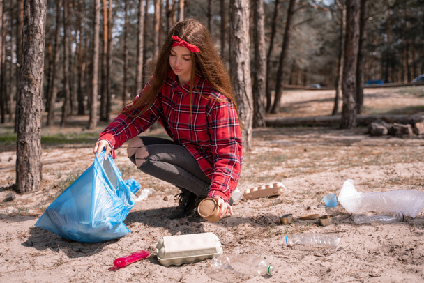 νεαρή γυναίκα που κρατάει σακούλα σκουπιδιών και μαζεύει σκουπίδια στο έδαφος στο δάσος - Φωτογραφία, εικόνα