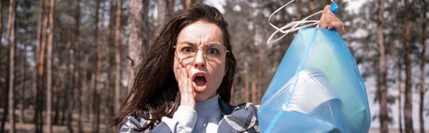 conmocionada joven mujer sosteniendo bolsa de basura azul en el bosque, pancarta - Foto, Imagen