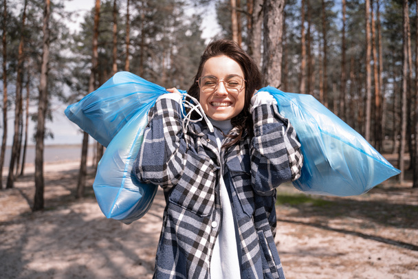 alegre joven en gafas sosteniendo bolsas de basura azul en el bosque  - Foto, Imagen