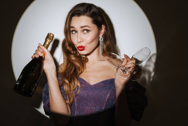 Beautiful excited young glamorous woman wearing cocktail dress and accessories posing in a spotlight, isolated background, holding champagne bottle and glasses, celebrating - Foto, Bild