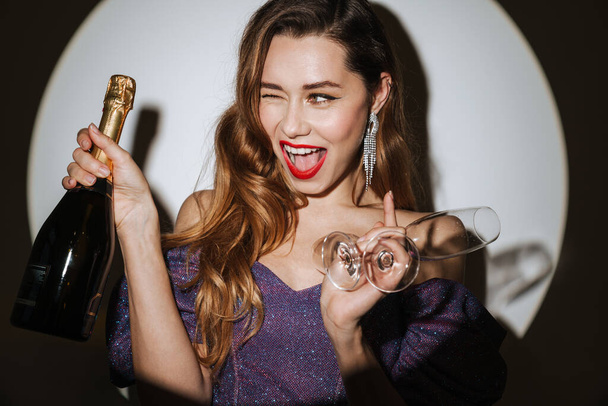 Beautiful excited young glamorous woman wearing cocktail dress and accessories posing in a spotlight, isolated background, holding champagne bottle and glasses, celebrating - Foto, immagini