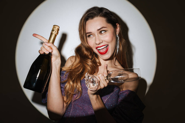 Beautiful excited young glamorous woman wearing cocktail dress and accessories posing in a spotlight, isolated background, holding champagne bottle and glasses, celebrating - Φωτογραφία, εικόνα