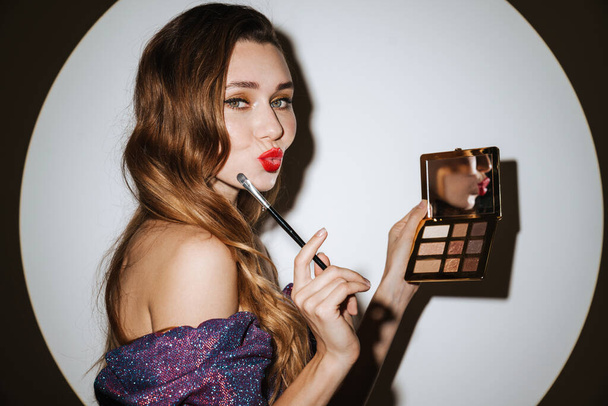 Beautiful young glamorous woman wearing cocktail dress and accessories posing in a spotlight, isolated background, getting ready for a party, applying makeup, holding eyeshadow palette - Foto, immagini