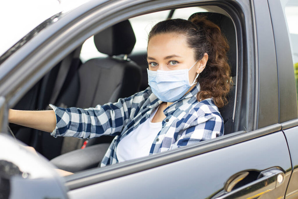 Young woman in a mask sitting in a car, protective mask against coronavirus, driver on a city street during a coronavirus outbreak. - Φωτογραφία, εικόνα