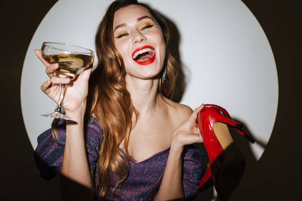 Beautiful excited young glamorous woman wearing cocktail dress and accessories posing in a spotlight, isolated background, holding glass with champagne, carrying high heels shoes - Foto, immagini