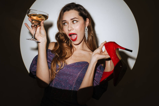Beautiful excited young glamorous woman wearing cocktail dress and accessories posing in a spotlight, isolated background, holding glass with champagne, carrying high heels shoes - Photo, Image