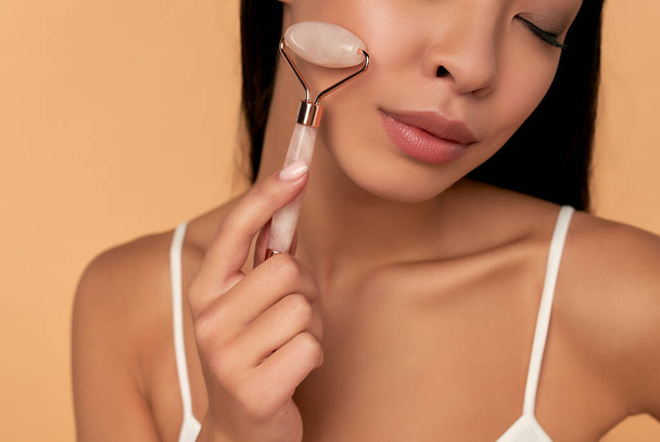透明感のある肌の若いアジア人女性がベージュ地にクオーツローラーで顔のマッサージをします。スパケア、フェイシャルスキンケア、美容整形. - 写真・画像