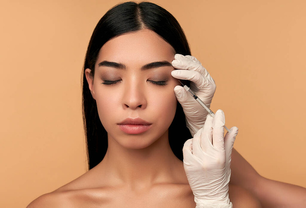 Young Asian woman with clean radiant skin gets botox injections for contour tightening, lip augmentation on a beige background. Spa care, facial skin care, beauty cosmetology. - Fotoğraf, Görsel