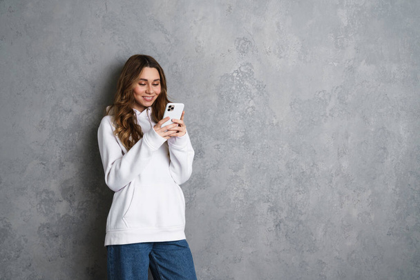 Happy young girl in casual wear using smartphone isolated on a gray background - Φωτογραφία, εικόνα