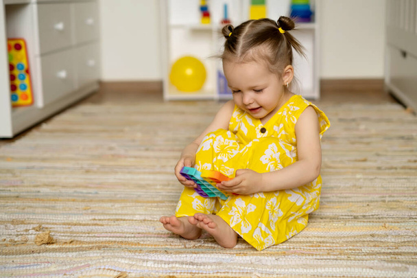 Ein Mädchen spielt auf dem Fußboden im Kinderzimmer mit dem beliebten Pop-it-Spielzeug. Ein Spielzeug zur Entwicklung der Beweglichkeit. Entwicklung der Feinmotorik. Ein Kind hält ein Popit-Spielzeug in den Händen. - Foto, Bild