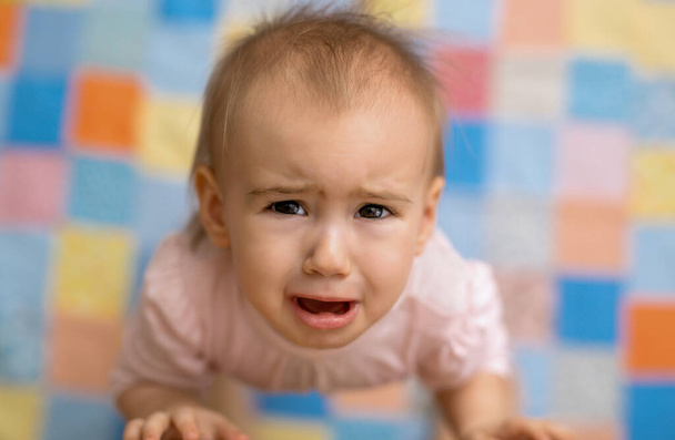 O bebê chora de pé ao lado de sua mãe e olhando para cima. Um bebé giro e choroso. Retrato de uma menina em lágrimas no fundo de um cobertor colorido. medo nos olhos da criança - Foto, Imagem