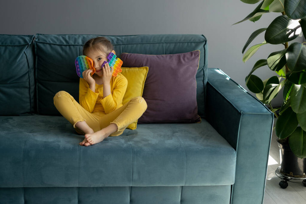 A fascinating sensory toy . Colorful and bright pop it toy. Simple dimple. The child is sitting on a sofa with yellow and purple pillows. A little girl is playing with a modern popit toy - Photo, Image