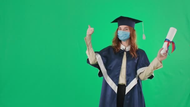 distancia de graduación, mujer joven en vestido y sombrero se alegra de recibir el diploma y muestra la clase con el pulgar hacia arriba mientras está de pie sobre fondo verde, croma clave - Imágenes, Vídeo