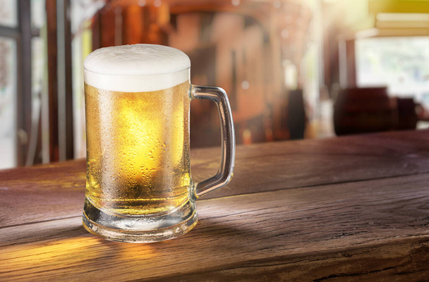 Cooled mug of pale beer with condensation drops on glass surface on the wooden table. Blurred bar at the background. - Фото, изображение