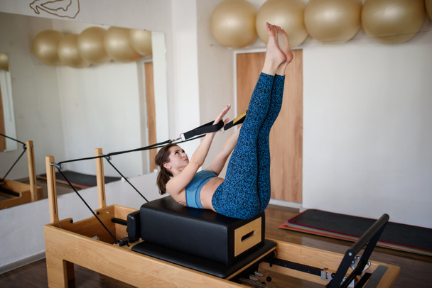 Una giovane donna in abiti sportivi fa Pilates su un riformatore alzando le gambe. - Foto, immagini