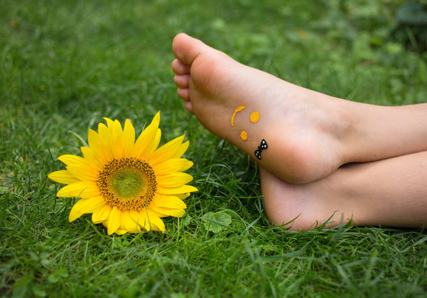sorriso sono disegnati sui piedi nudi di un bambino sdraiato sull'erba. Un fiore di girasole giallo giace nelle vicinanze sull'erba. gioia, allegra atmosfera positiva, infanzia felice. Ciao estate, energia della natura - Foto, immagini