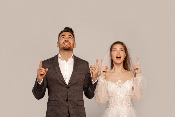 astonished bride and groom looking up and pointing with fingers isolated on grey - Foto, imagen