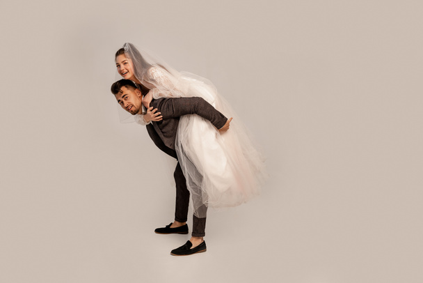 full length view of cheerful bride piggybacking on groom isolated on grey - Zdjęcie, obraz