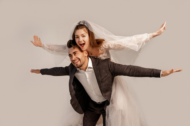fröhliche Braut huckepack auf glücklichen Bräutigam isoliert auf grau - Foto, Bild