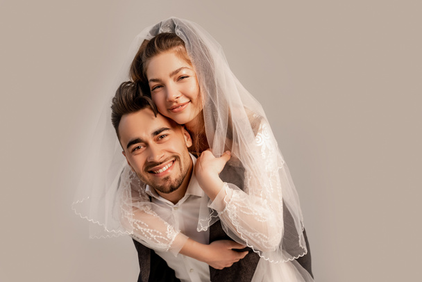 Lächelnde Braut huckepack auf glücklichem Bräutigam isoliert auf grau mit Fliederton - Foto, Bild