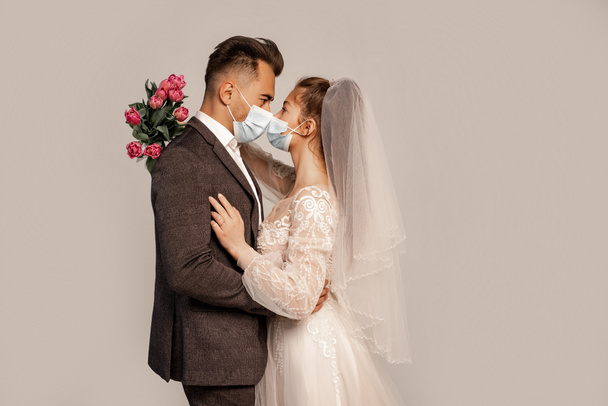 side view of bride and groom in protective masks standing face to face isolated on grey - Foto, afbeelding