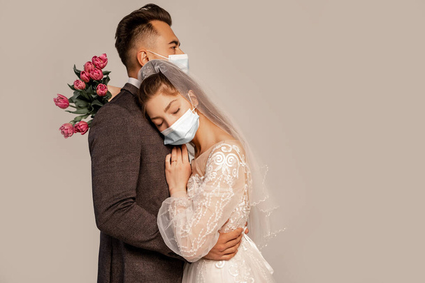 man in medical mask embracing bride leaning on him with closed eyes isolated on grey - Foto, afbeelding