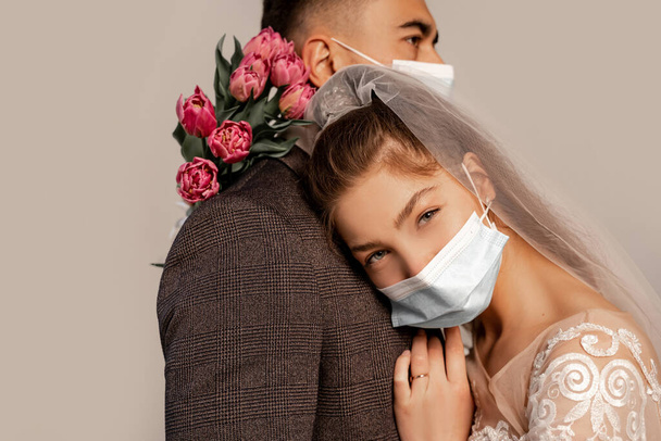 young bride in medical mask leaning on chest of groom and looking at camera isolated on grey - Φωτογραφία, εικόνα