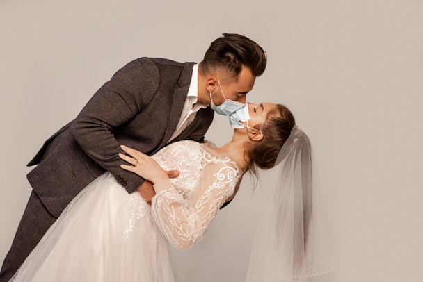 man in medical masks kissing elegant bride isolated on grey with lilac shade - 写真・画像
